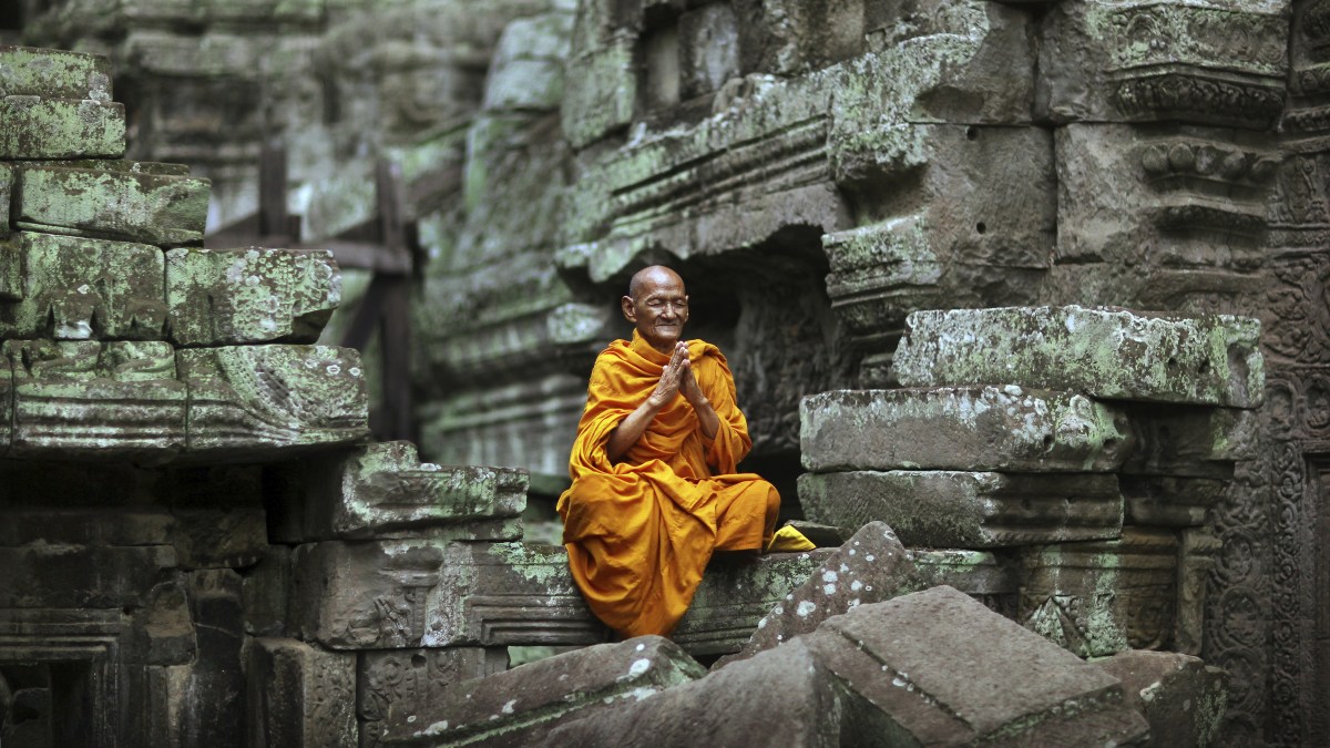 Hands held together in prayer, a Buddhist monk elder wearing bright orange robes meditates in the lotus position amid the temple ruins at Ta Prohm temple in Cambodia. (Timothy Allen/Creative RM/Getty Images)