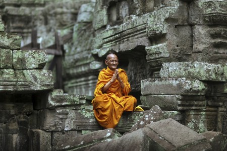 Hands held together in prayer, a Buddhist monk elder wearing bright orange robes meditates in the lotus position amid the temple ruins at Ta Prohm temple in Cambodia. (Timothy Allen/Creative RM/Getty Images)