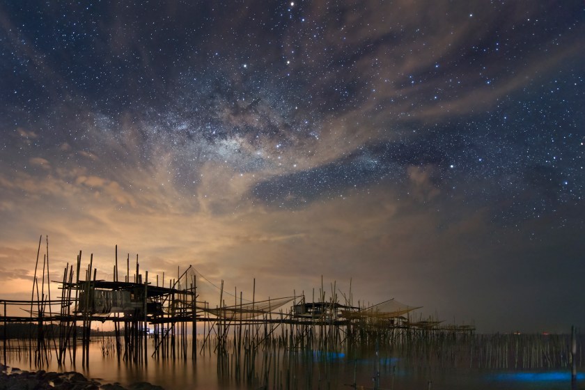 The natural light of the Milky Way battles with the light pollution over the fishing village, or kelong, in Batu Pahat, Malaysia. In the lower right hand corner, there is also bioluminescence in the waters at the bottom of the kelong. (Yuyu Wang)