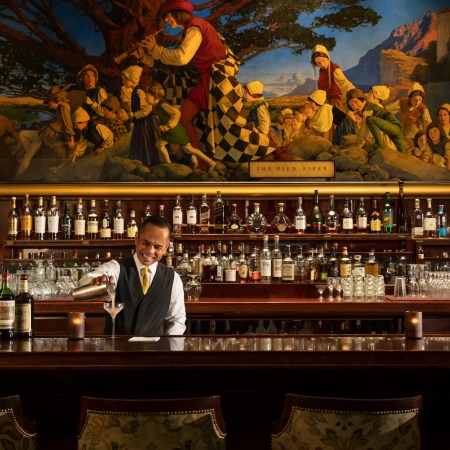 Bartender pouring a cocktail from a shaker into a glass on a dark wooden bar in front of shelves with bottles and a large art print on the wall