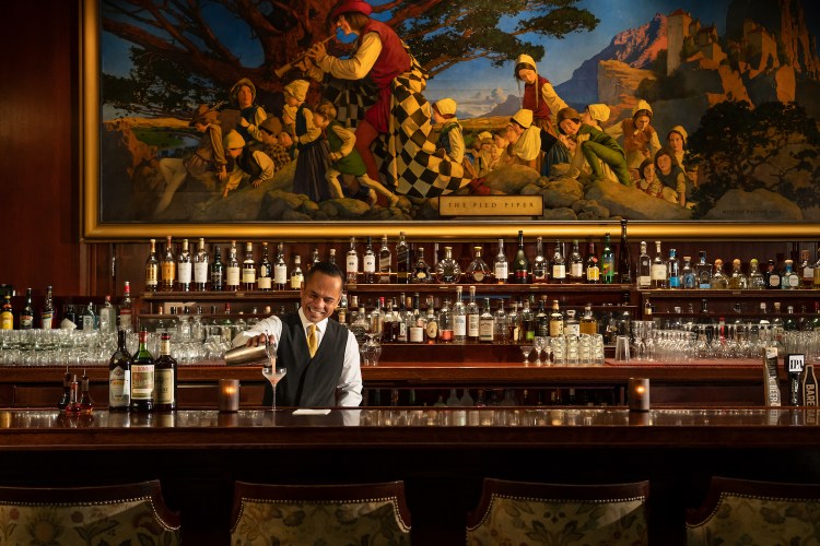Bartender pouring a cocktail from a shaker into a glass on a dark wooden bar in front of shelves with bottles and a large art print on the wall