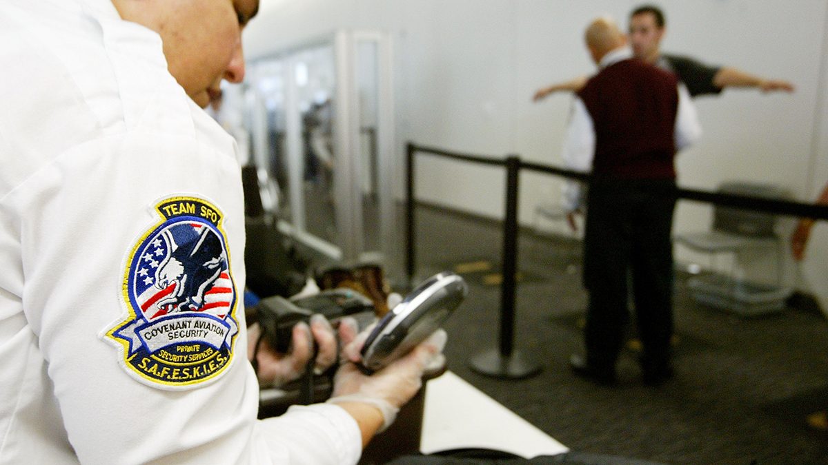 A Transportation Security Administration baggage screener inspects an electronics device as a man is screened in the background at the international terminal of San Francisco International Airport August 5, 2003 in San Francisco, California. The TSA told screeners today to pay close attention to cameras, laptop computers and cell phones, addressing a concern that terrorists could attempt to hide explosives in electronic devices.  (Justin Sullivan/Getty Images)