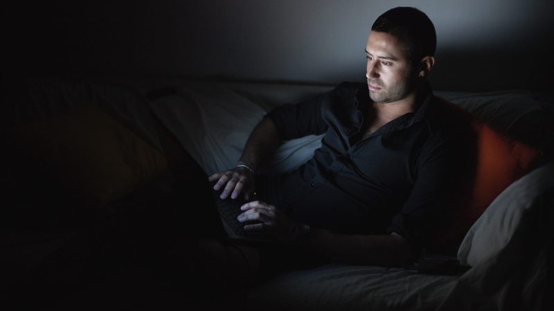 Man lying on sofa and working on his laptop at night.