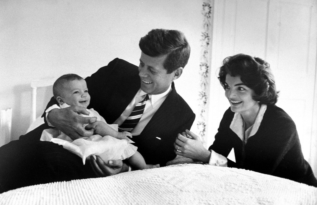 John F. Kennedy cuddling his darling baby daughter Caroline who is smiling as her mom Jackie looks on in delight while relaxing on bed at home.