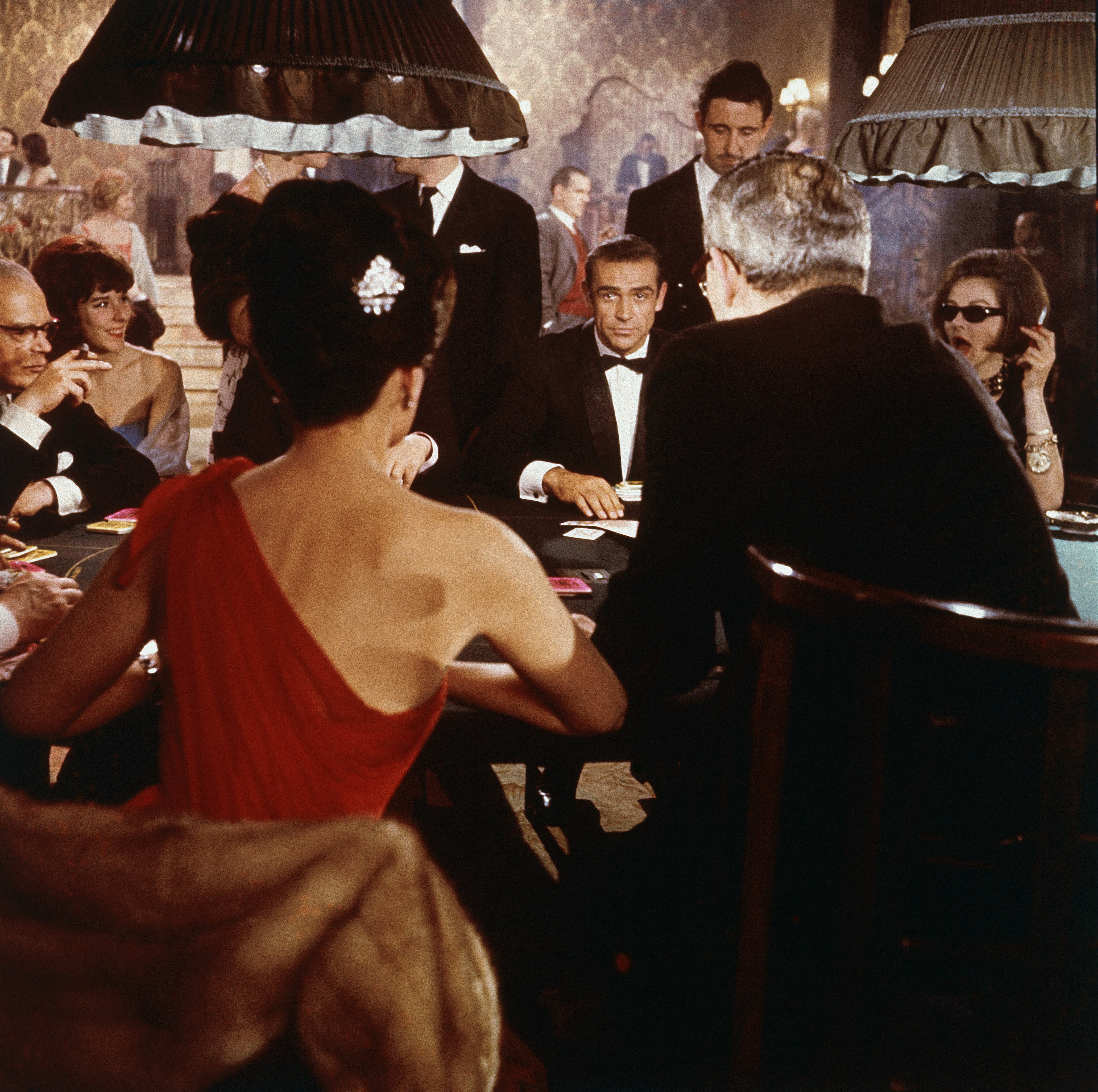 Scottish actor Sean Connery (center) as fictional secret agent James Bond sits at a casino card table in a scene from the film 'Dr. No,' directed by Terence Young, 1962. British actress Eunice Gayson sits with her back to the camera in a red, off the shoulder dress. (MGM Studios/Courtesy of Getty Images)
