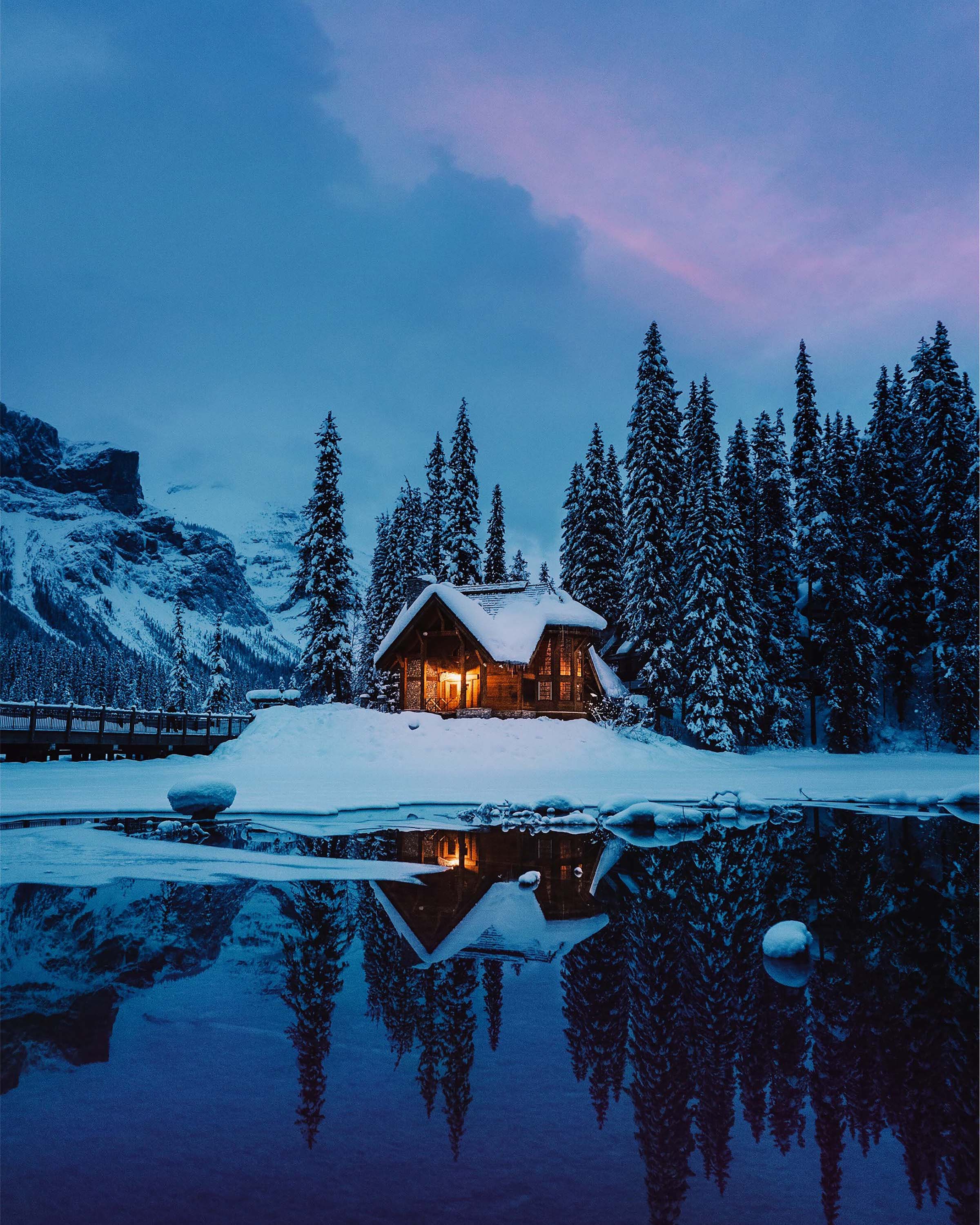 A cozy log cabin in the middle of an snowy forest with a lake in the foreground