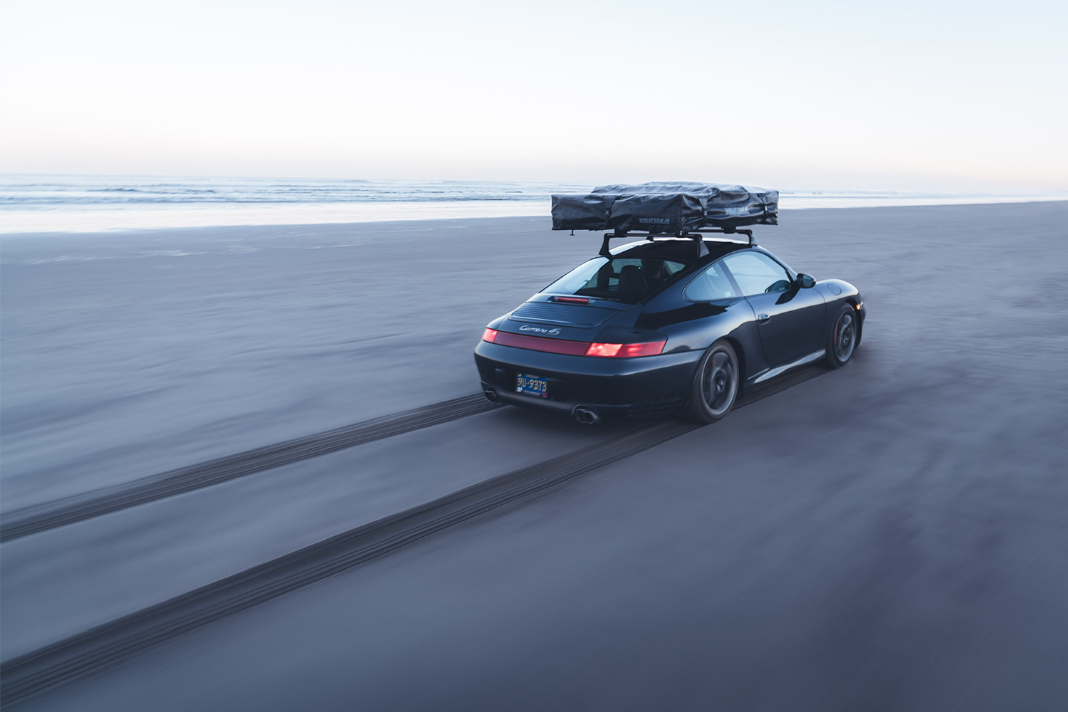 Brock Keen's black Porsche 996 911 with a rooftop tent speeding down the sandy beach next to the ocean