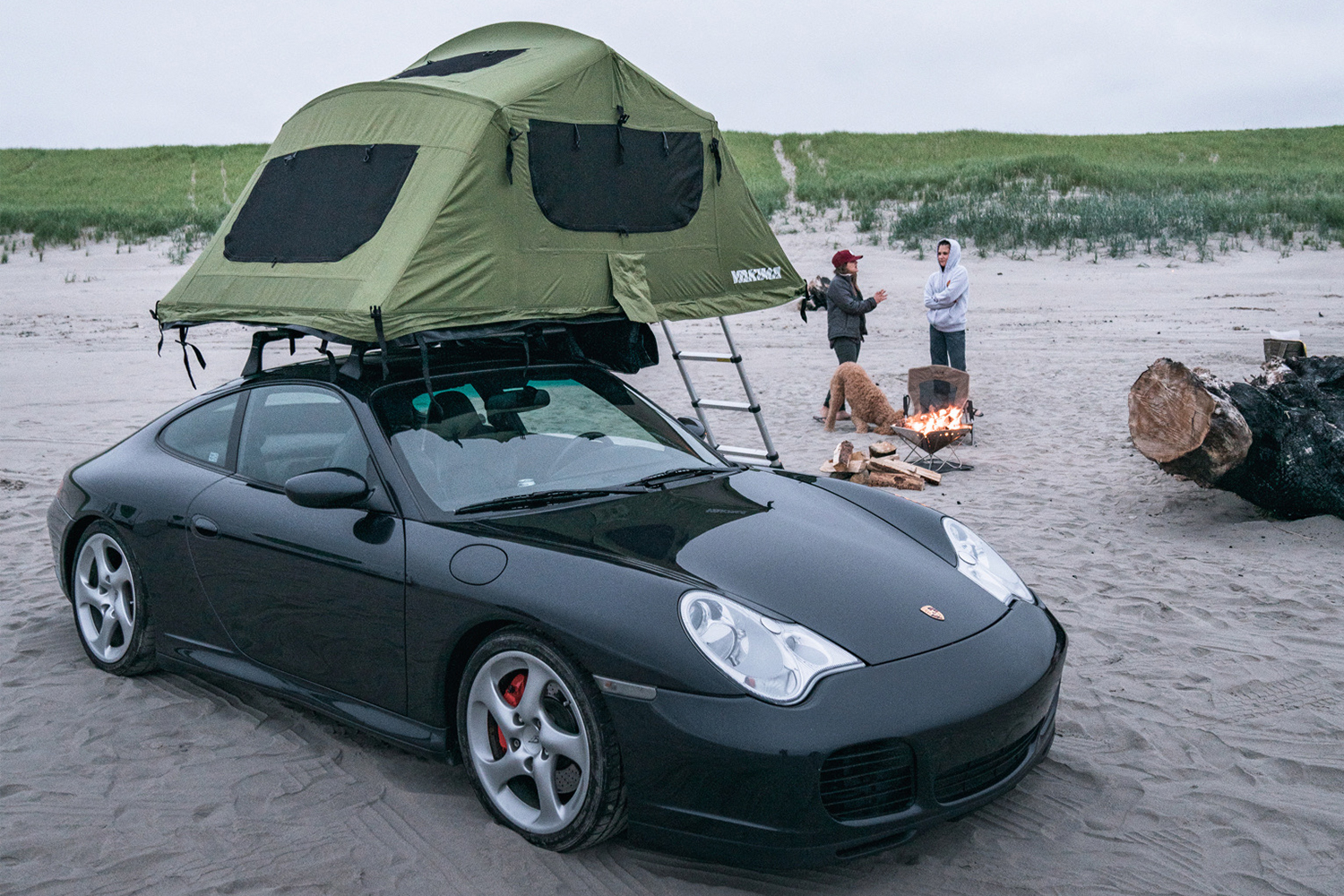 A black 2004 Porsche 996 C4S, or Porsche 911, with a green Yakima rooftop tent owned by Brock Keen of Instagram handle @996roadtrip sitting on a beach with two women in the background next to a campfire