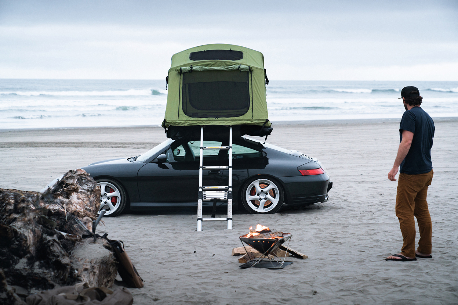 A black Porsche 911 with a green Yakima rooftop car camping tent with a ladder coming down the side sitting next to the ocean on a beach while owner Brock Keen of Instagram account @996roadtrip stands to the right
