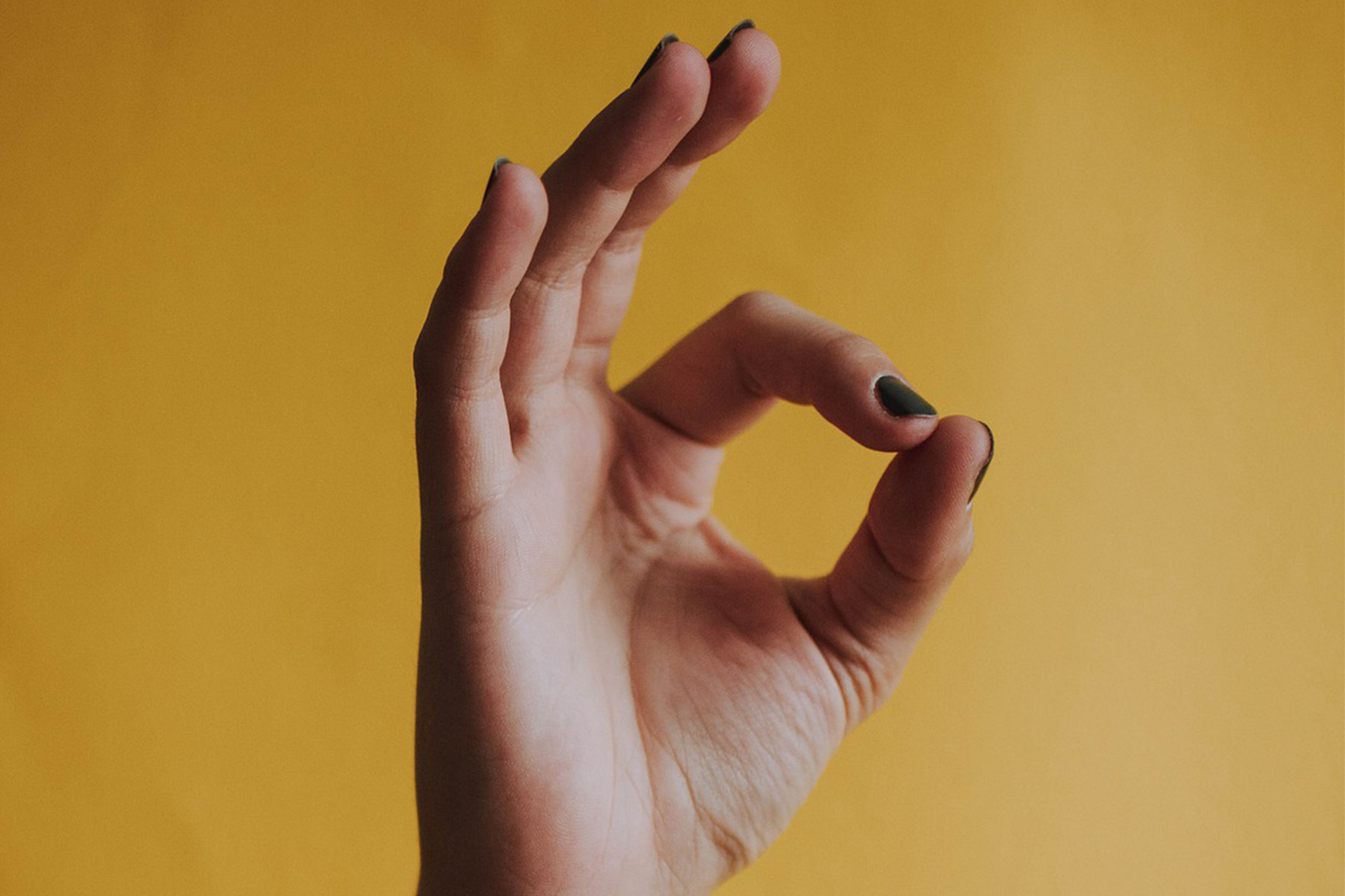 White hand in O-K gesture, with dark green painted fingernails, on yellow background