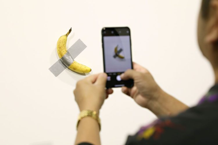 People post in front of Maurizio Cattelan's "Comedian" presented by Perrotin Gallery and on view at Art Basel Miami 2019 at Miami Beach Convention Center on December 6, 2019 in Miami Beach, Florida. Two of the three editions of the piece, which feature a banana duct-taped to a wall, have reportedly sold for $120,000.  (Photo by Cindy Ord/Getty Images)