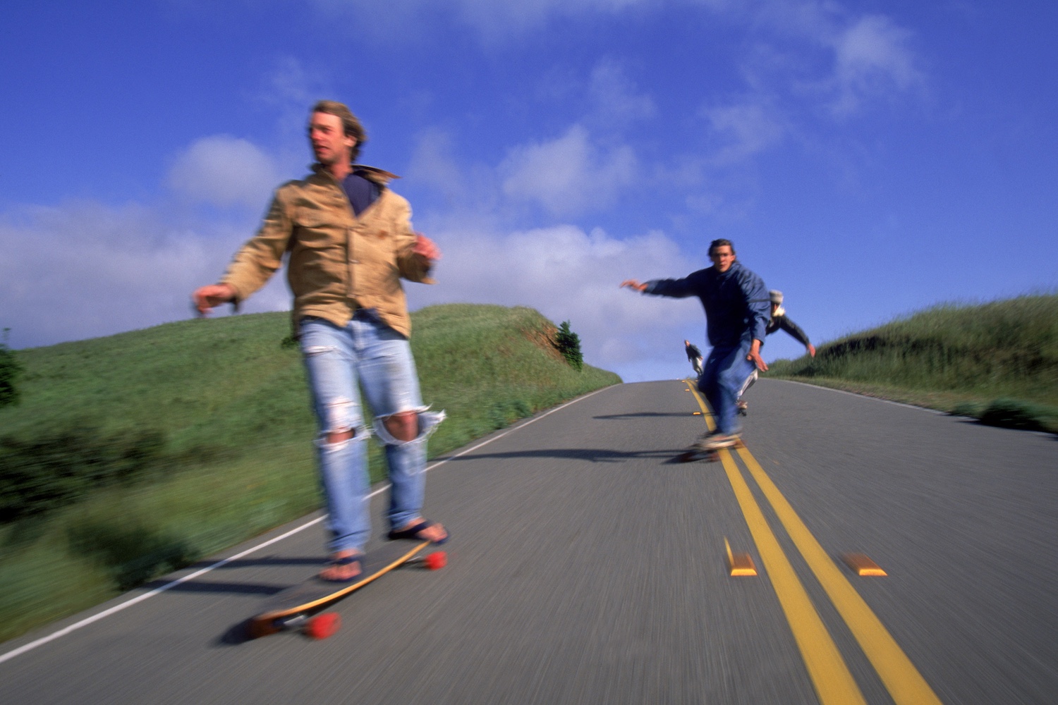Three longboarders riding down a scenic hill on a sunny day, with motion blur emphasizing speed.