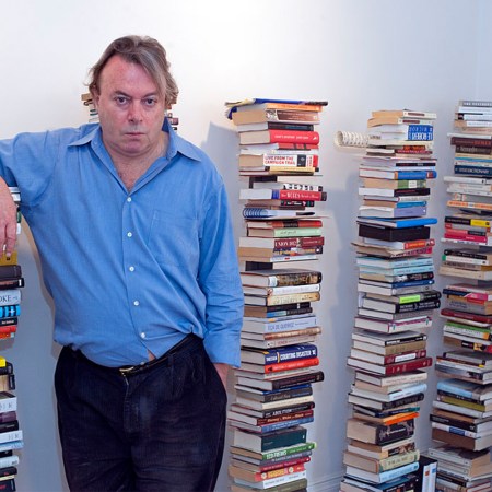 Christopher Hitchens standing next to piles of books