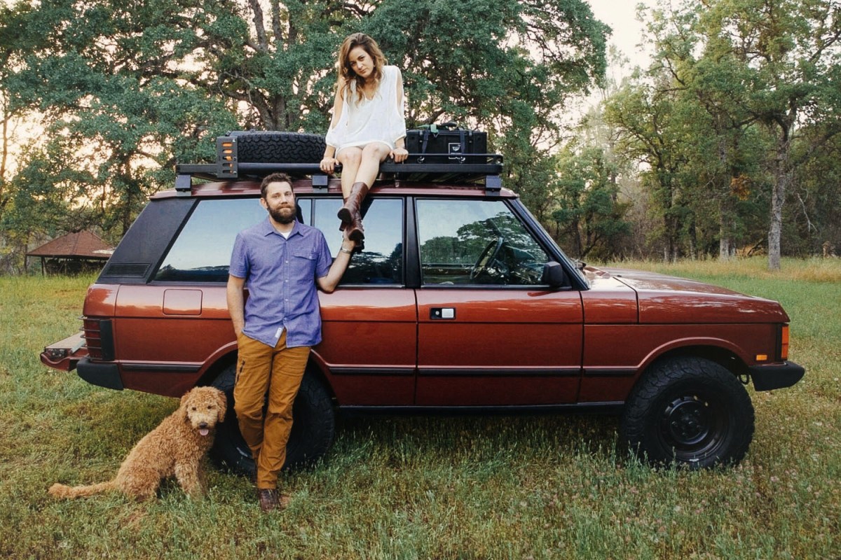 A man, woman and goldendoodle dog next to a red 1991 Range Rover Classic SUV