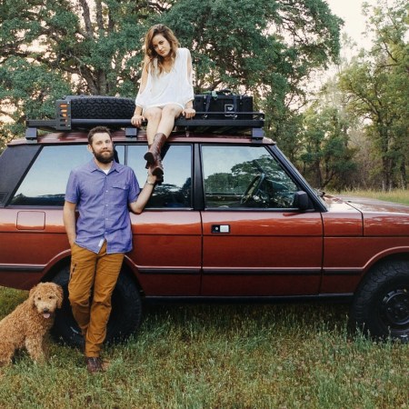 A man, woman and goldendoodle dog next to a red 1991 Range Rover Classic SUV