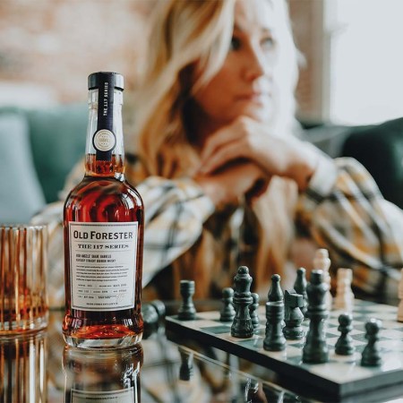 Old Forester Master Taster Jackie Zykan sitting at a table with a chess set and a bottle and glass of Old Forester 117 High Angels' Share