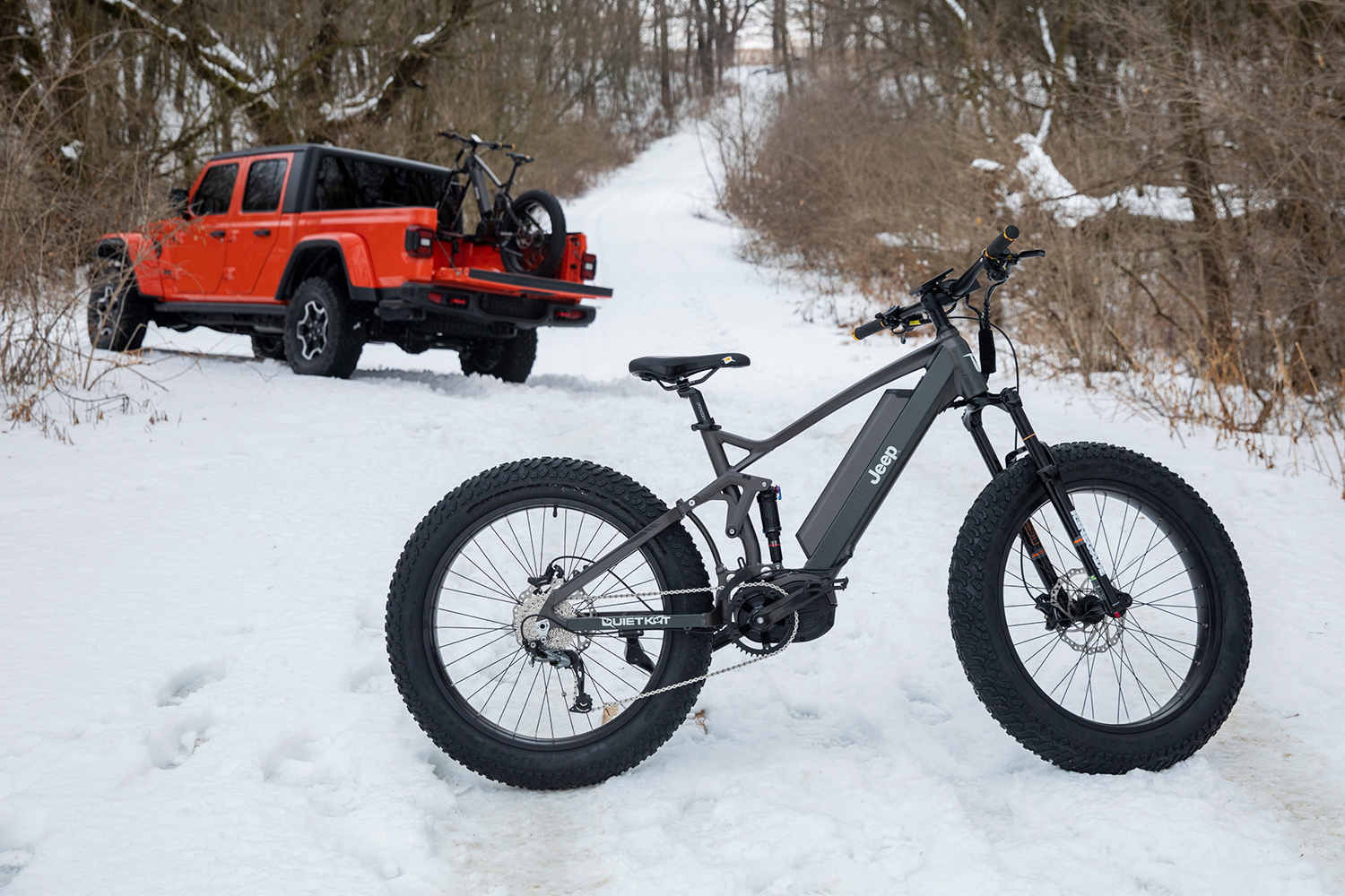 A Jeep e-bike sitting in the snow with an orange Gladiator pickup truck in the background