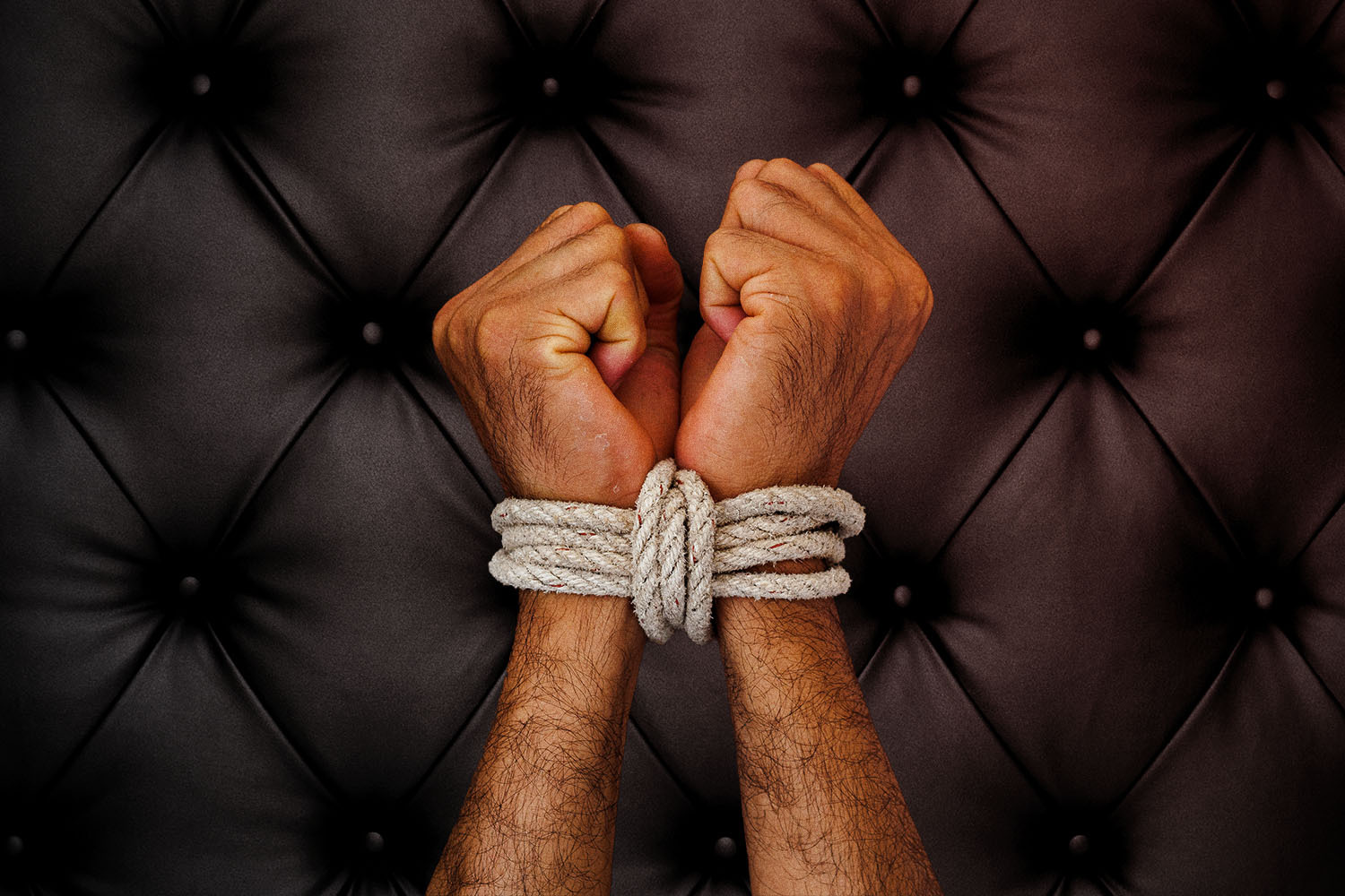 Man's wrists tied with white rope against black leather background.