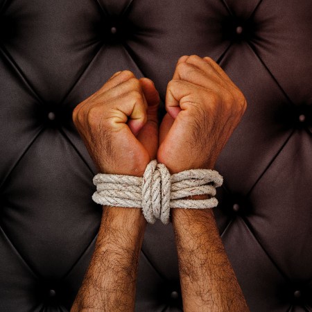 Man's wrists tied with white rope against black leather background.