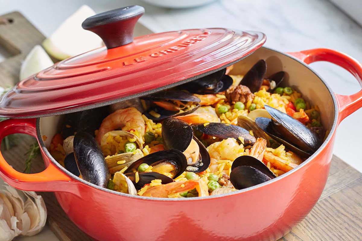 A red Le Creuset sauteuse pan with food on the stove. Some colors of this pan are up to $120 off at Sur La Table.