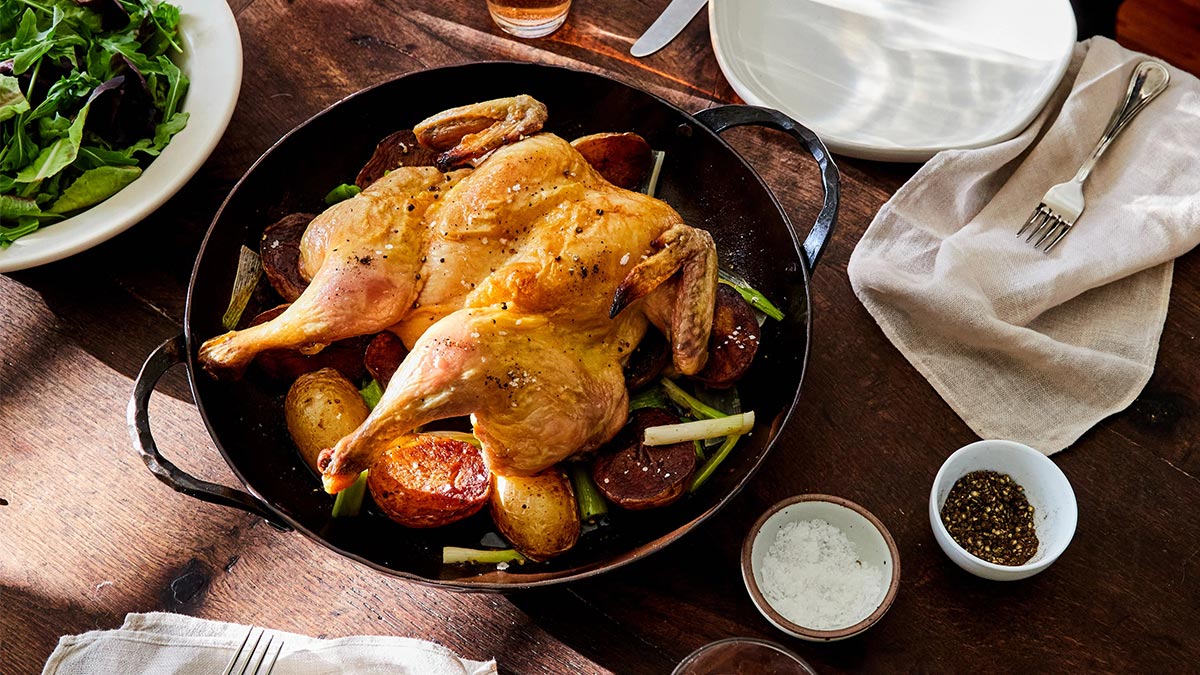 The Smithey Carbon Steel Round Roaster sitting on a table with a chicken and potatoes in it