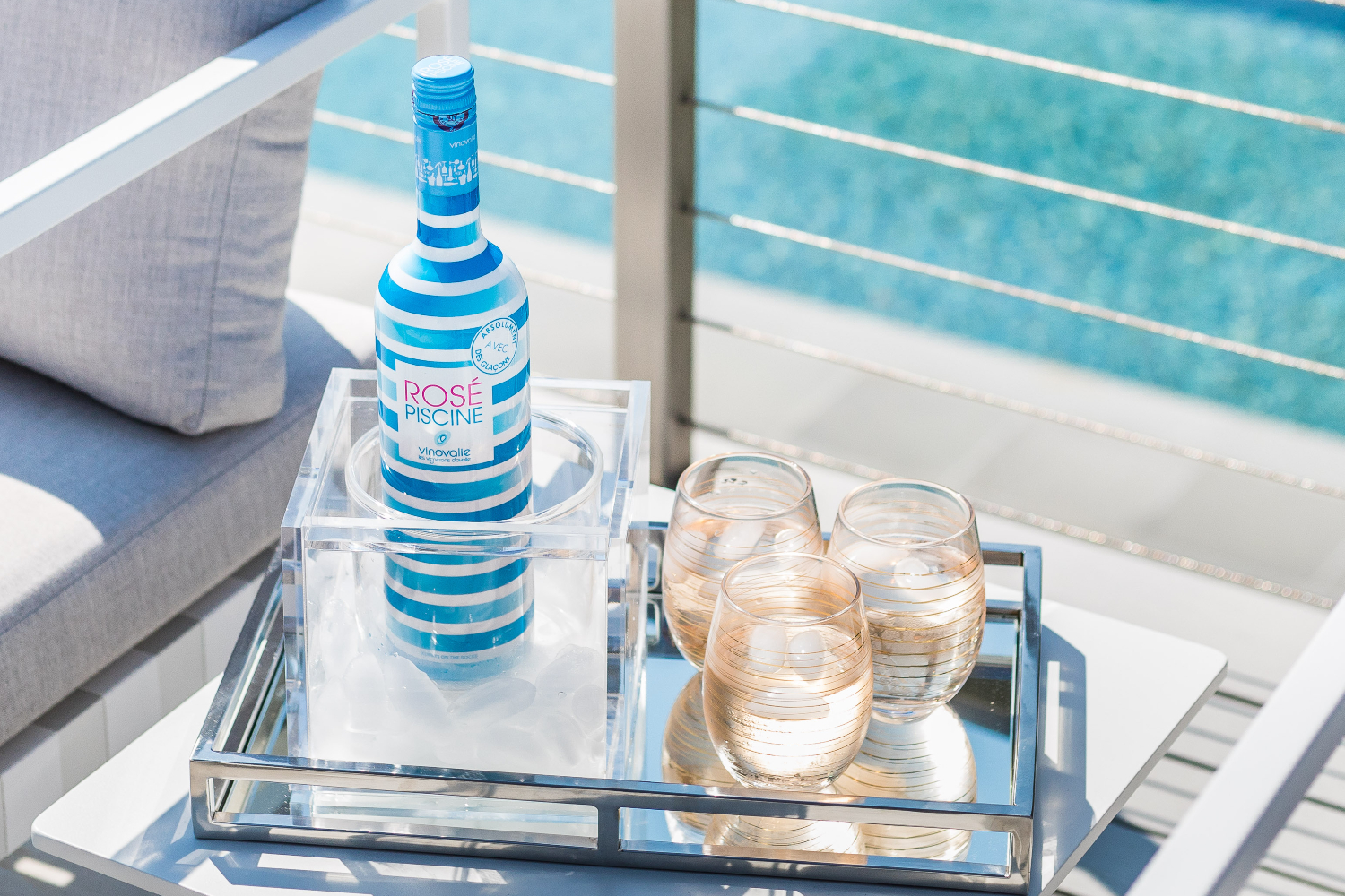 Blue-striped bottle of Rosé Piscine on a tray next to three stemless glasses of rosé with blue pool water in the background.