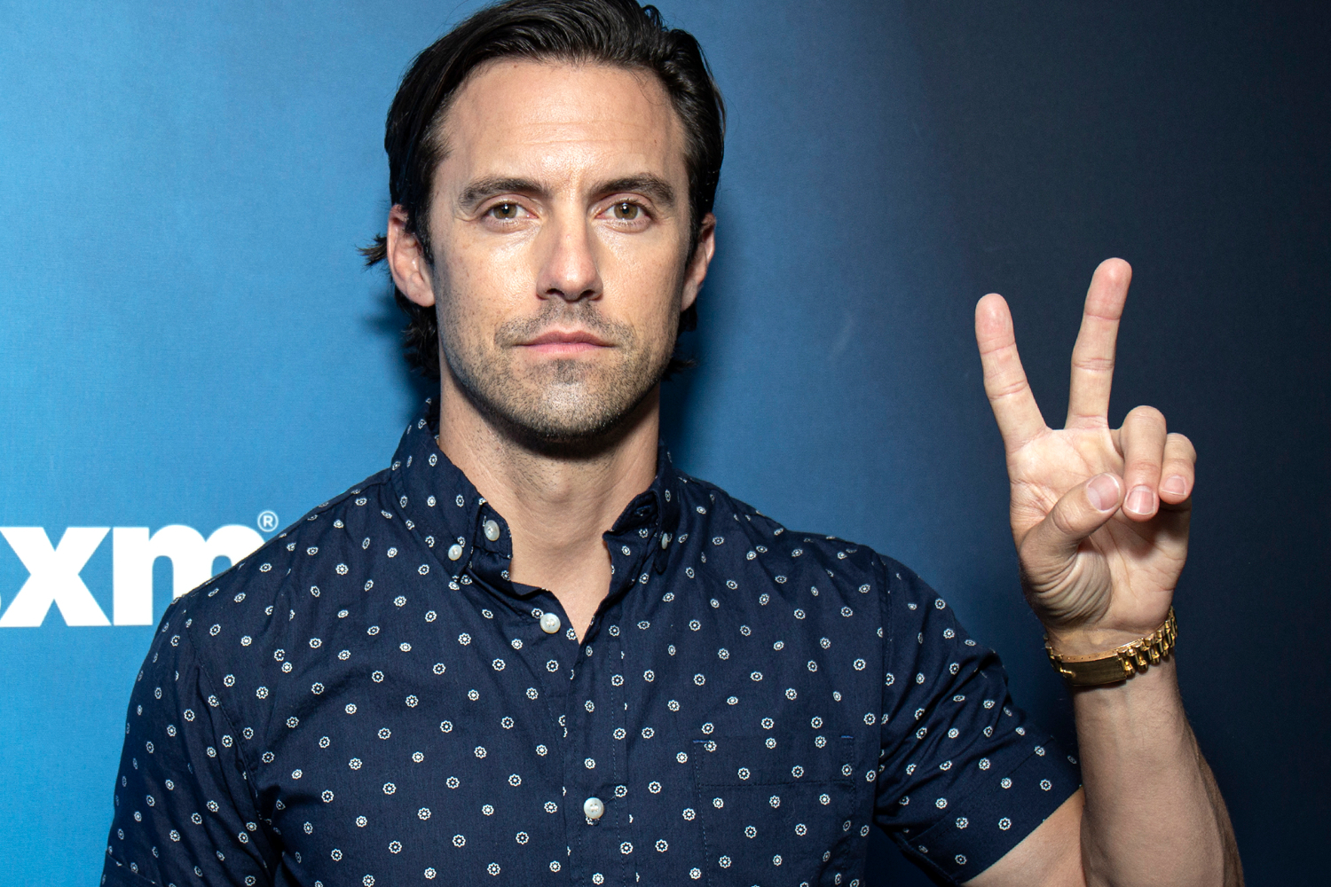 Milo Ventimiglia throws up a peace sign while posing against a blue backdrop