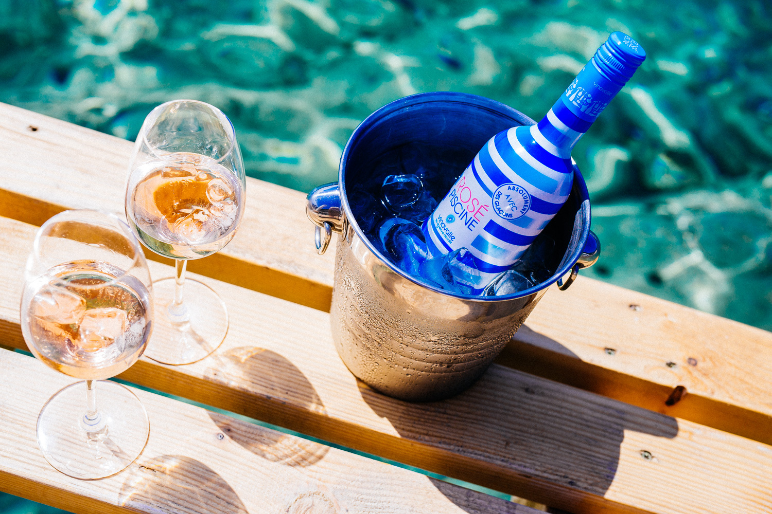 Blue and white striped bottle of Rosé Piscine in an ice bucket, next to two glasses of rosé, by blue-green water