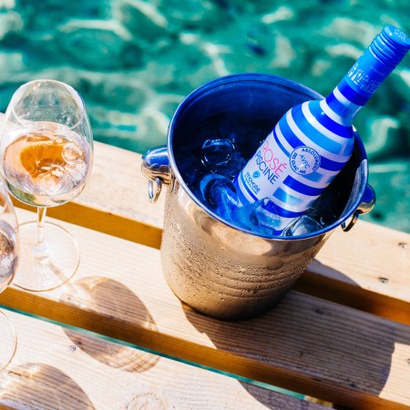 Blue and white striped bottle of Rosé Piscine in an ice bucket, next to two glasses of rosé, by blue-green water