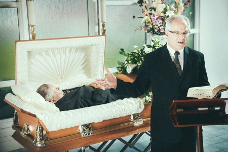 An older man giving a eulogy for another man (deceased) behind him in a coffin