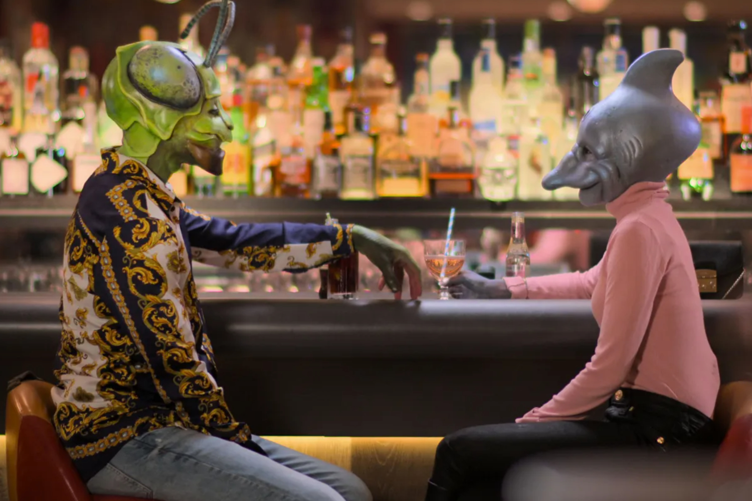A woman wearing a dolphin head and a man wearing a bug head sit at a bar in the trailer for Netflix's "Sexy Beasts."