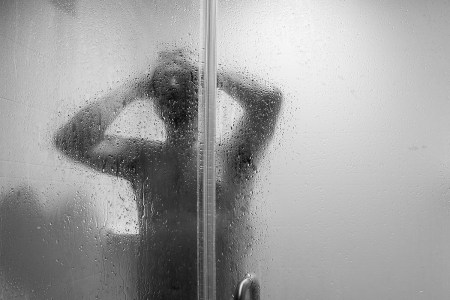 A black and white photo of a person showering, as soon through a shower door