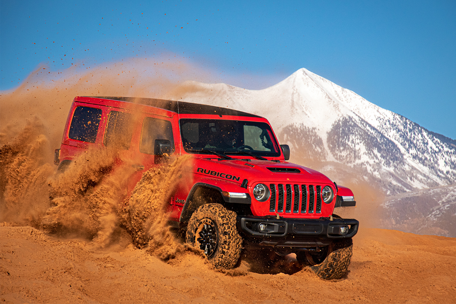 A red Jeep Wrangler Rubicon 392, the first-ever Wrangler with a V8 engine, churning up sand dunes in front of a snow-capped mountain