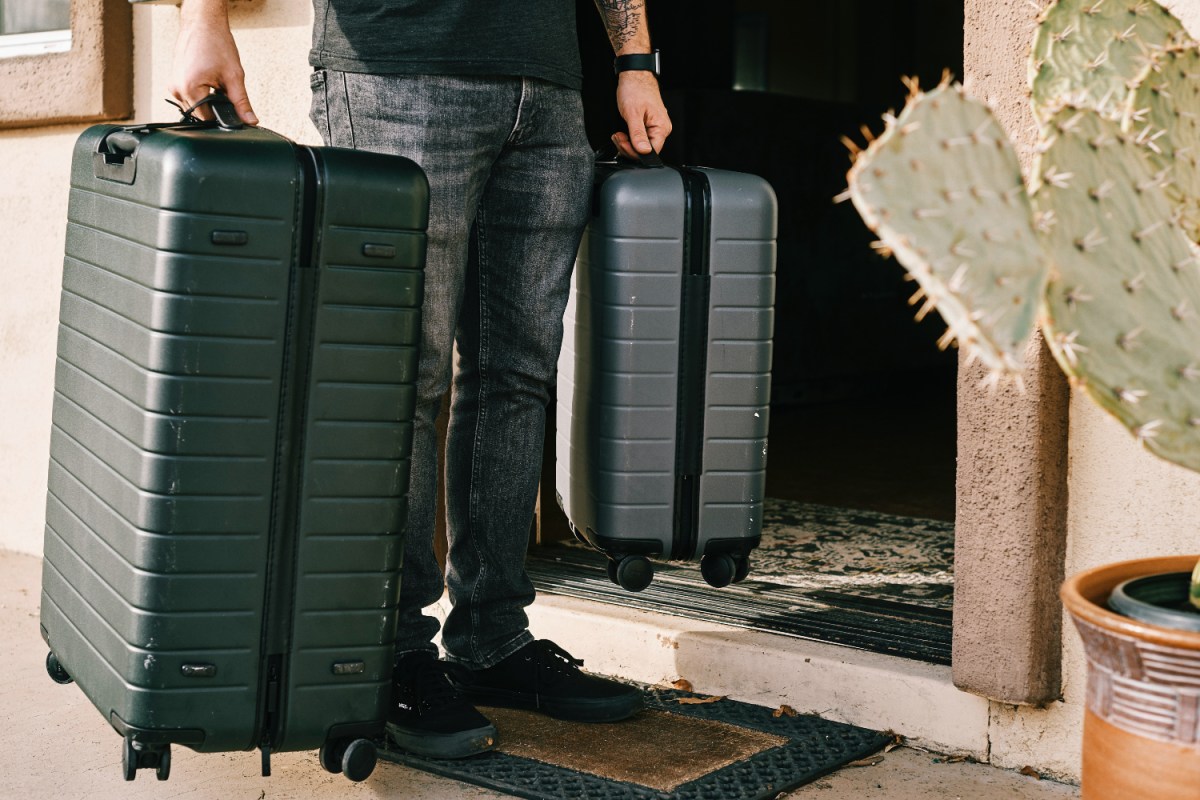 Man in jeans and black shirt stands near a door holding two rolling suitcases