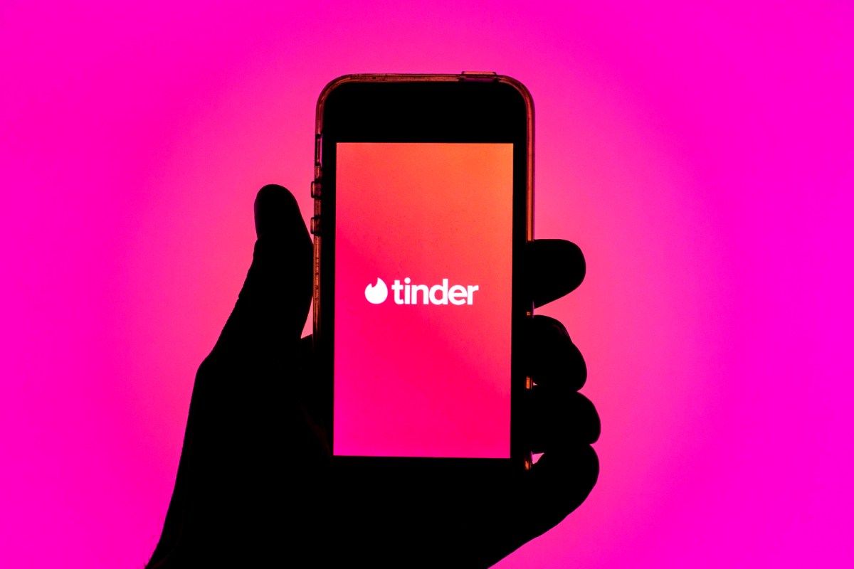 A silhouette of a hand holds a smartphone, displaying the Tinder logo against a pink background.