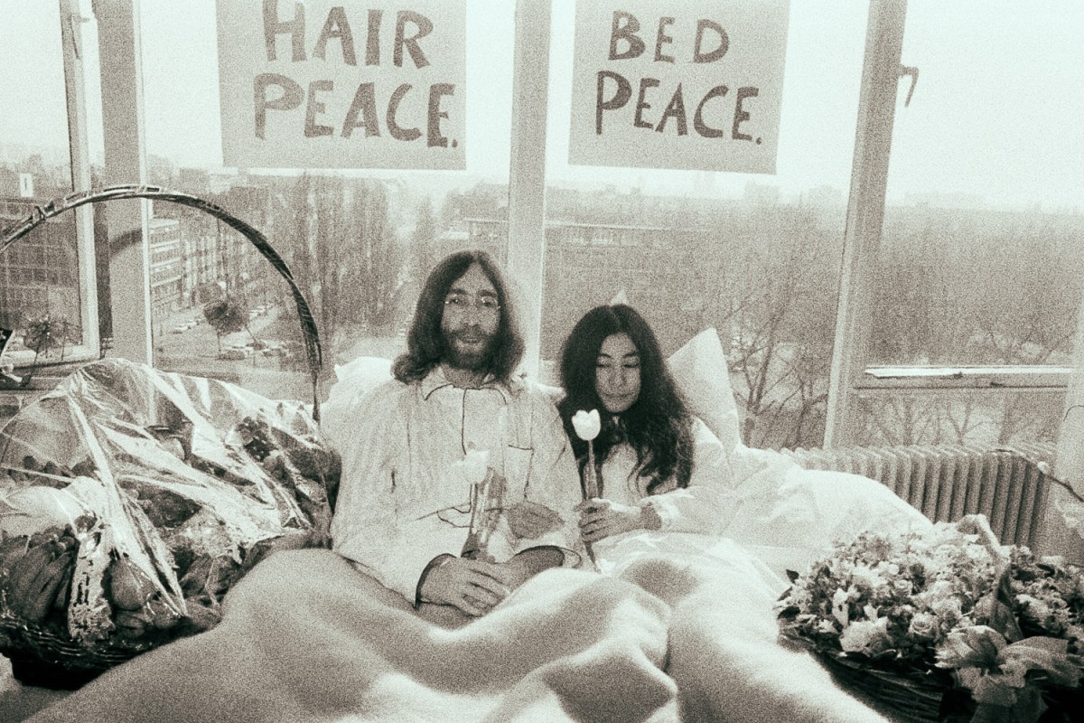 Black and white photo shows two hippies in bed together beneath signs reading "HAIR PEACE" and "BED PEACE."