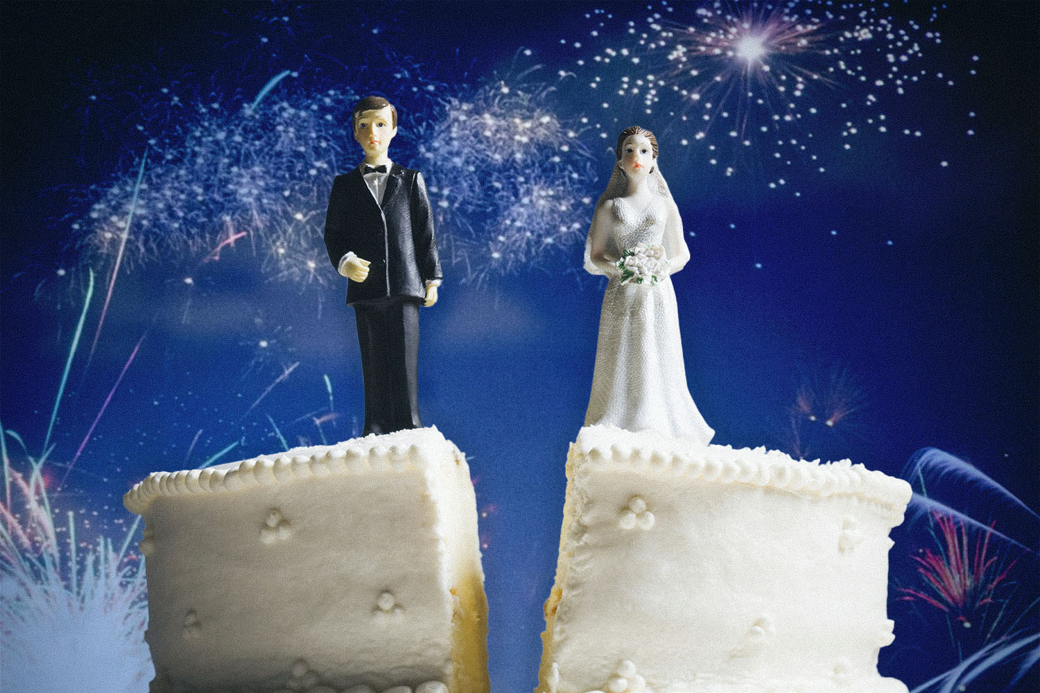 wedding cake split down the center, bride and groom cake toppers stand on either side. fireworks in the background.
