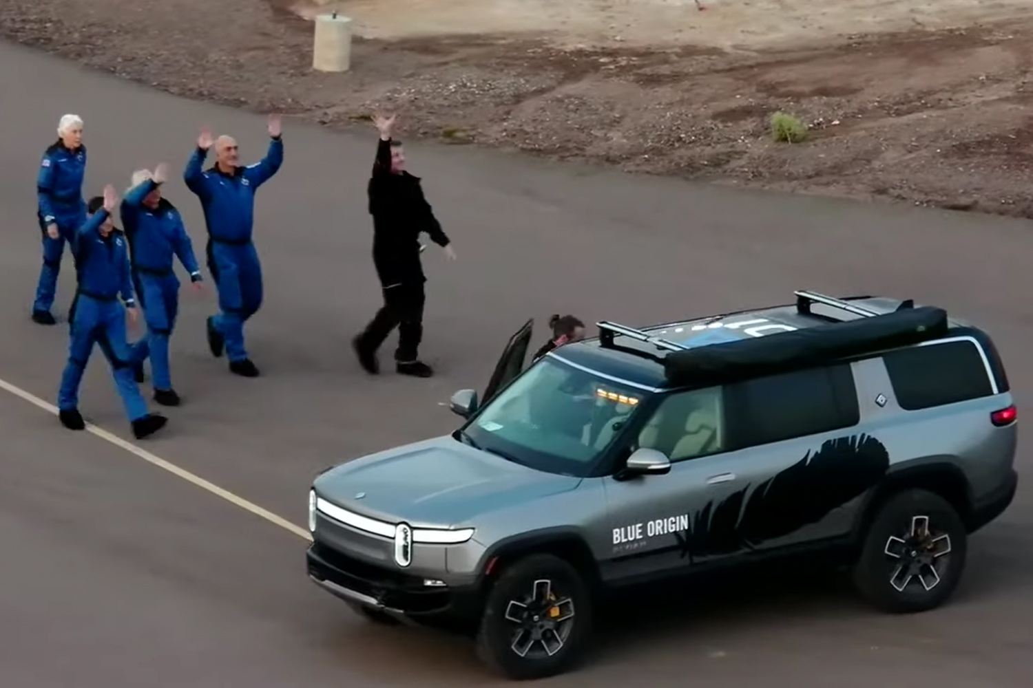 Jeff Bezos, Mark Bezos, Wally Funk and Oliver Daemen waving at the site of the Blue Origin rocket launch next to a Rivian SUV electric adventure vehicle