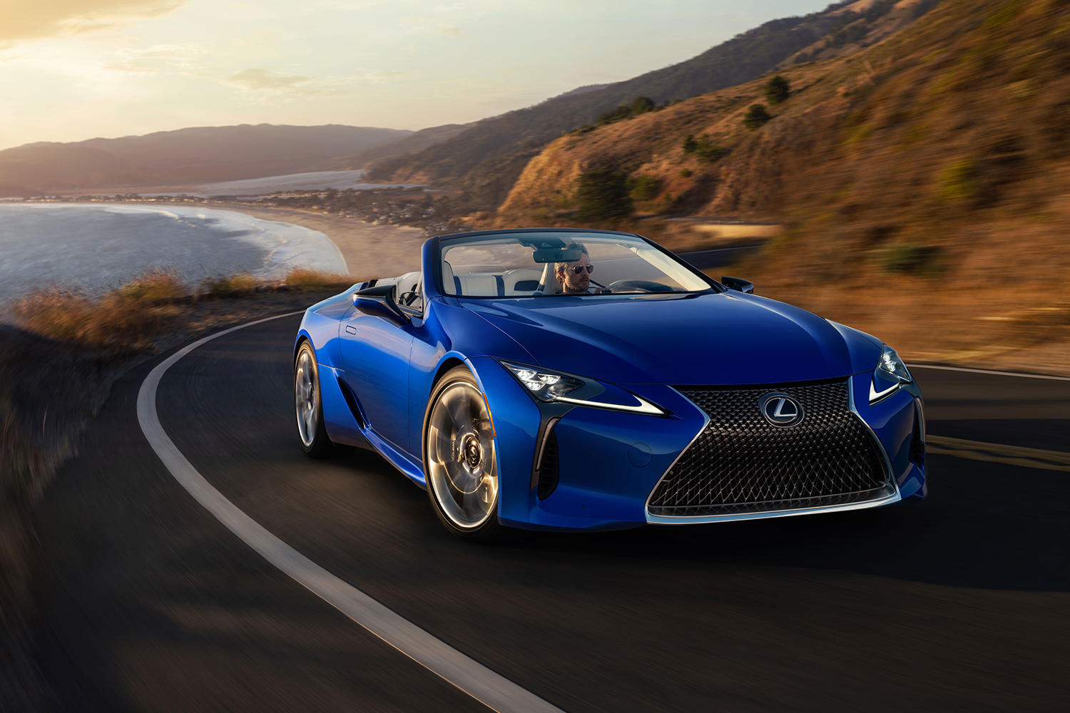 A man driving a blue 2021 Lexus LC 500 Convertible down a coastal road. The luxury grand tourer has become one of our favorites after test driving.