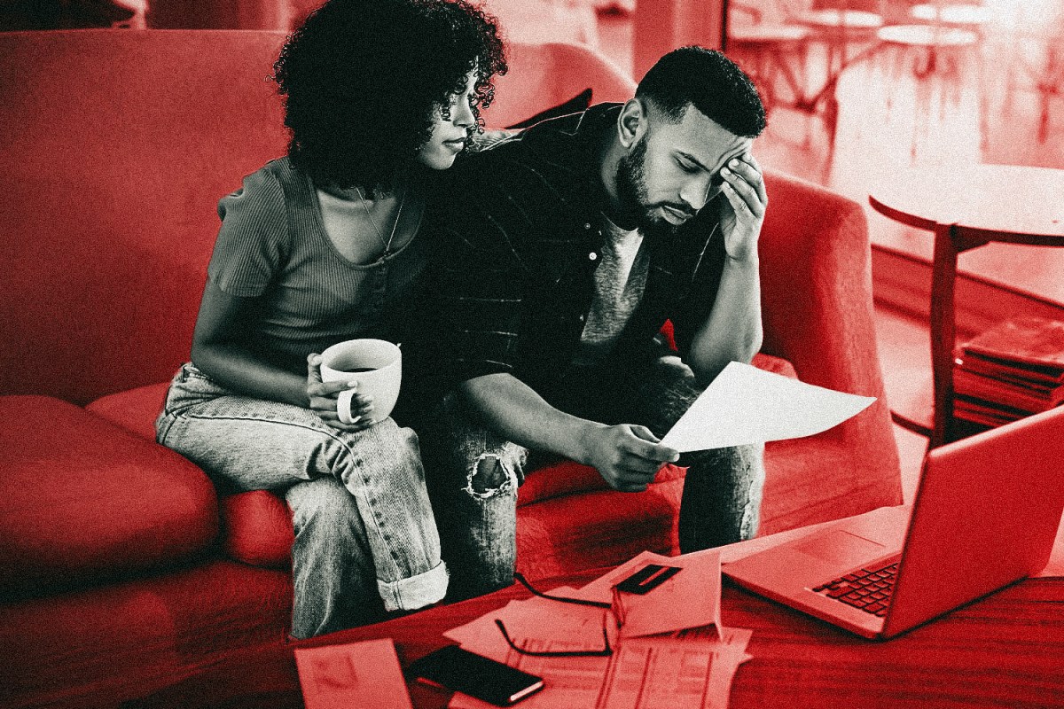 Financial infidelity occurs when a partner in a relationship lies or hides their financial circumstances from a partner. This photo shows a couple sitting on a couch together looking distressed while the man looks over financial documents.
