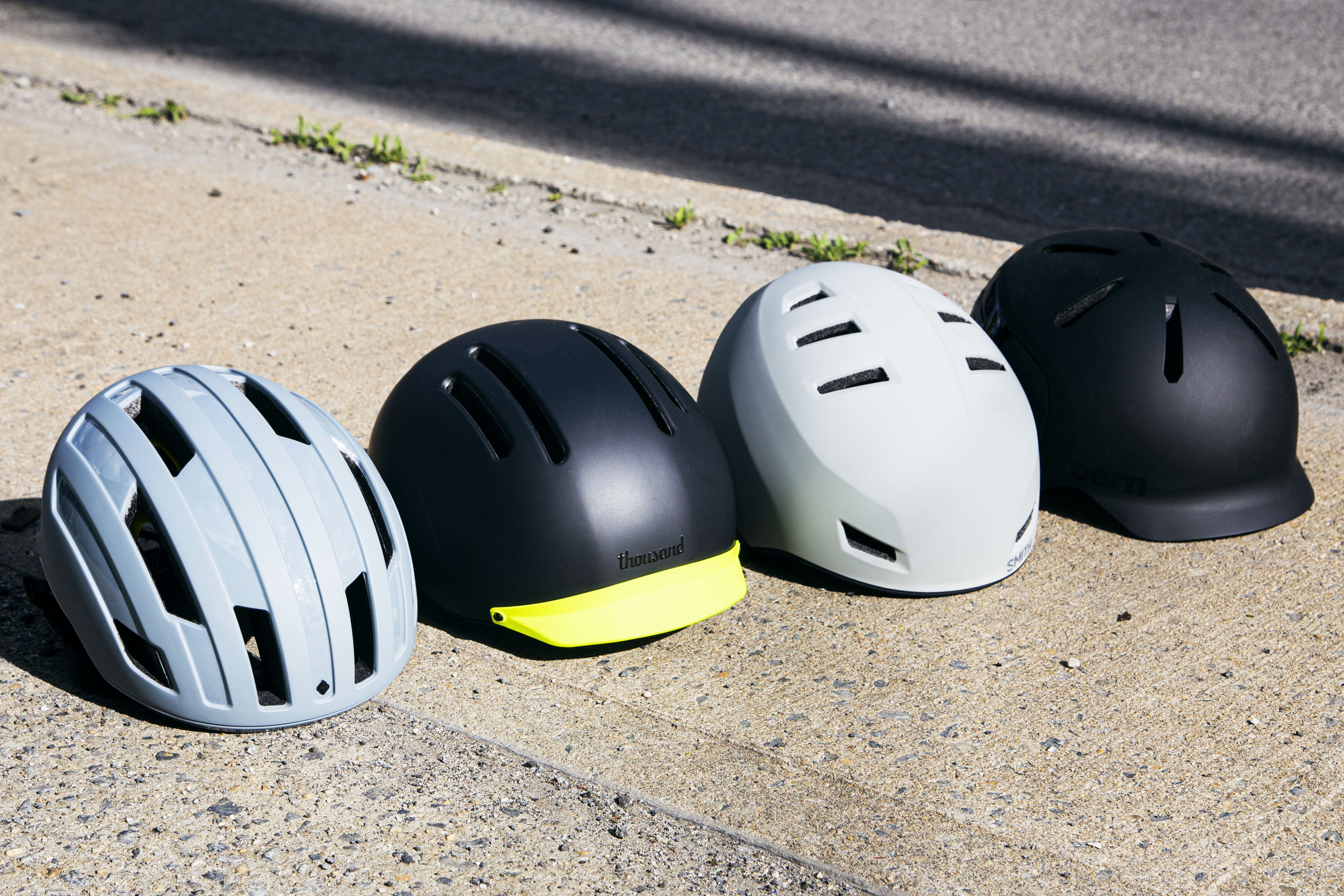 Four stylish, modern bicycle helmets — by Sweet Protection, Thousand, Smith and Bern, respectively — situated in a row on the sidewalk