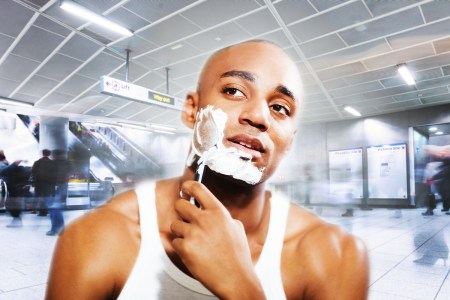A young man in an undershirt shaving his face with a razor inside a busy airport terminal