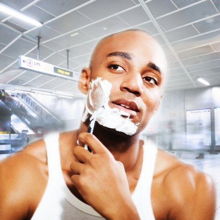 A young man in an undershirt shaving his face with a razor inside a busy airport terminal