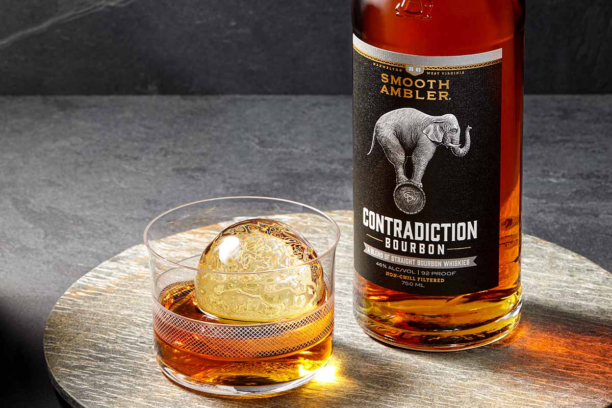 A bottle of Contradiction bourbon and a glass of whiskey with a ball of ice