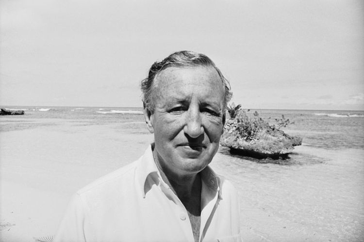 A black and white photo of author Ian Fleming on a beach. We take a look at how his wartime espionage work influenced writing his James Bond books.