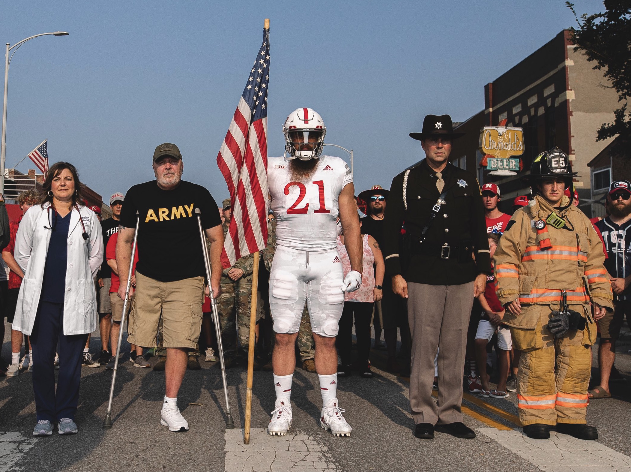 a university of nebraska football player holding a flag, flanked on both sides by US servicemen and front line emergency workers