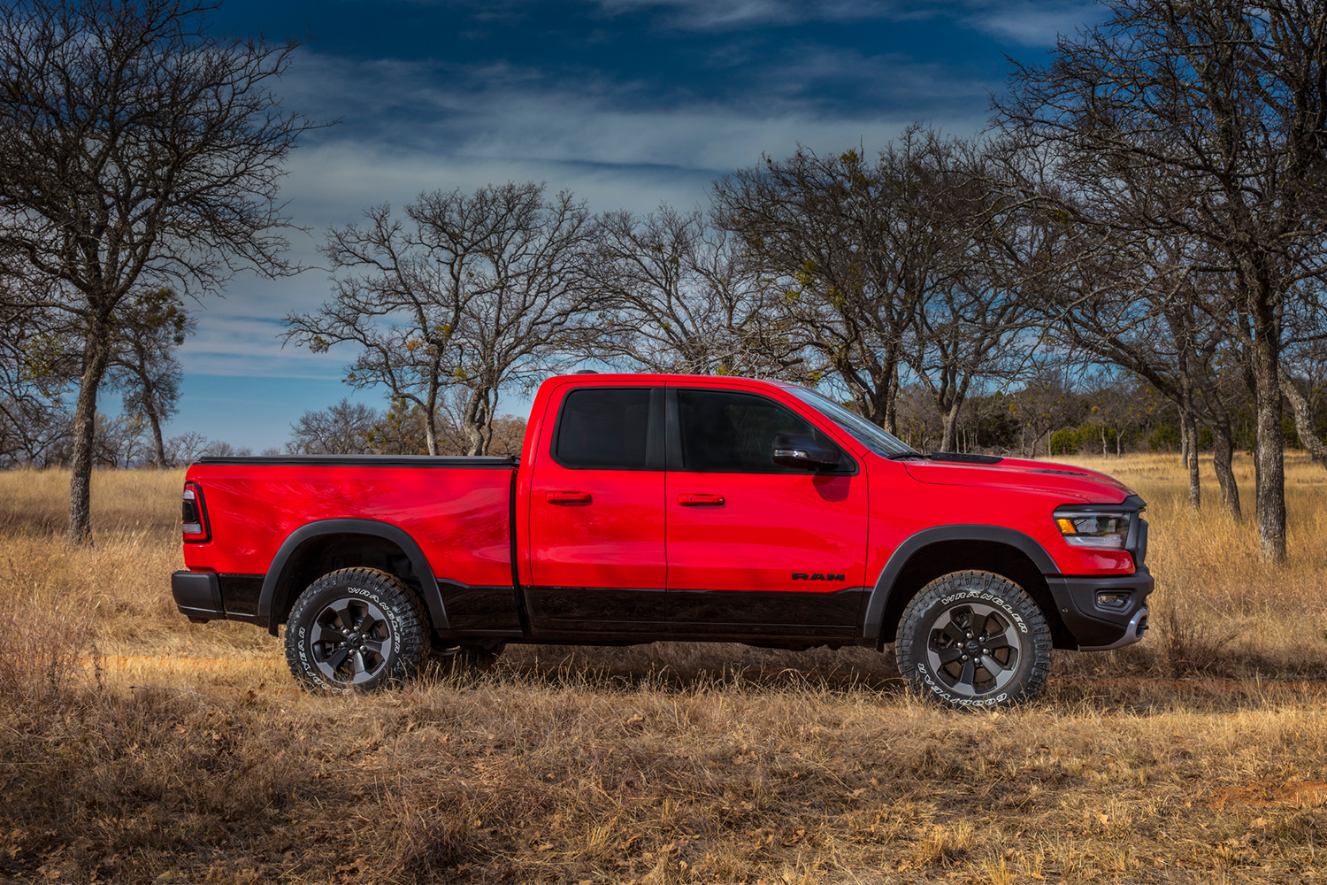 A profile view of a red Ram Trucks 1500 Rebel pickup sitting in the grass in front of a line of trees