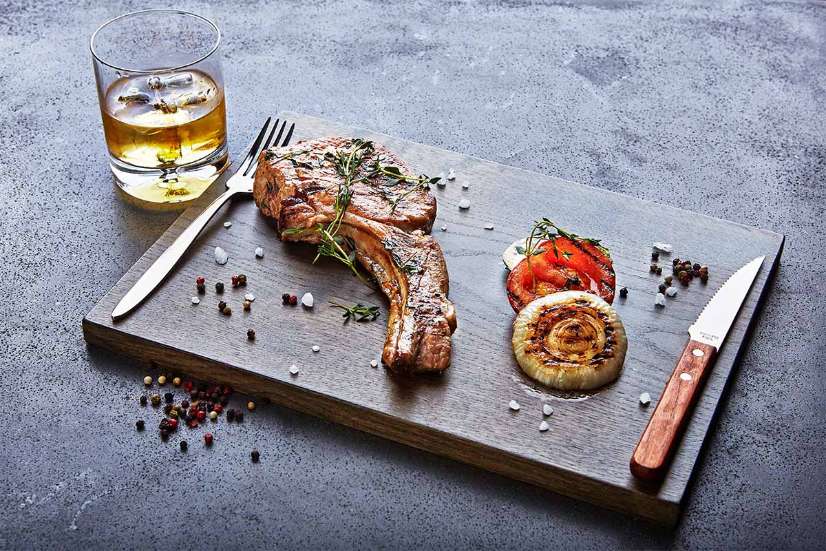 A piece of steak on a tray with a whiskey to the side. Besides combining with food pairings, whiskey is also good for cooking.