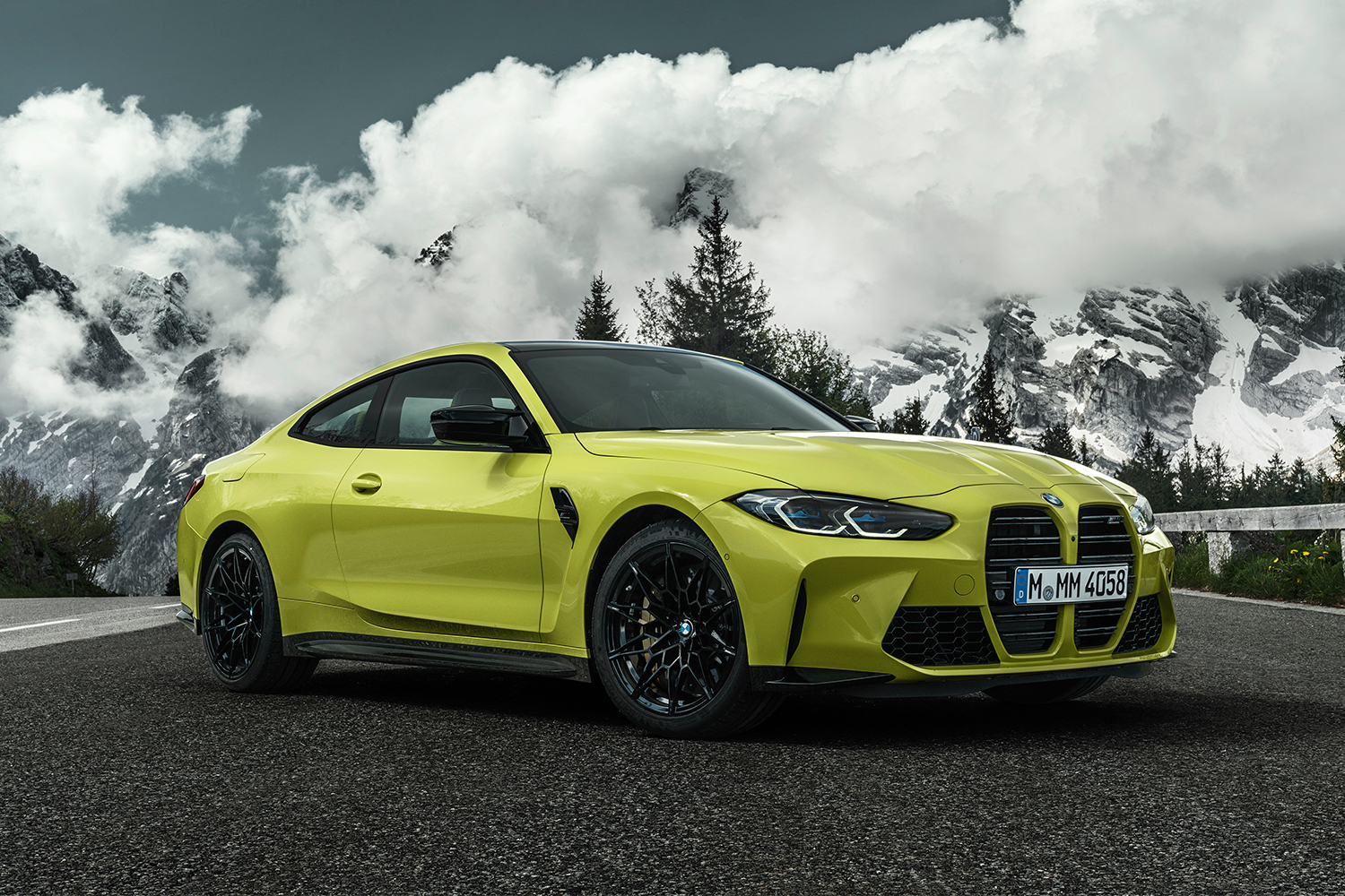The new BMW M4 Competition Coupé in yellow sits still on the road with snowy mountains and trees at the cloud line in the background