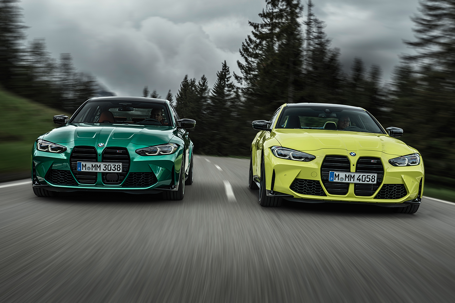 A green 2021 BMW M3 Competition Sedan on the left driving fast next to a 2021 BMW M4 Competition Coupe in yellow on the right with trees looming in the background